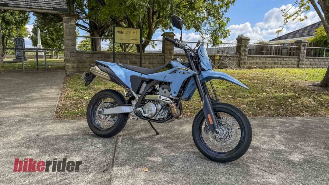Blue Suzuki dual-sport motorcycle parked on a concrete driveway with a stone wall, trees, and a clear sky in the background. The word 'bikerider' appears on the ground near the front wheel.