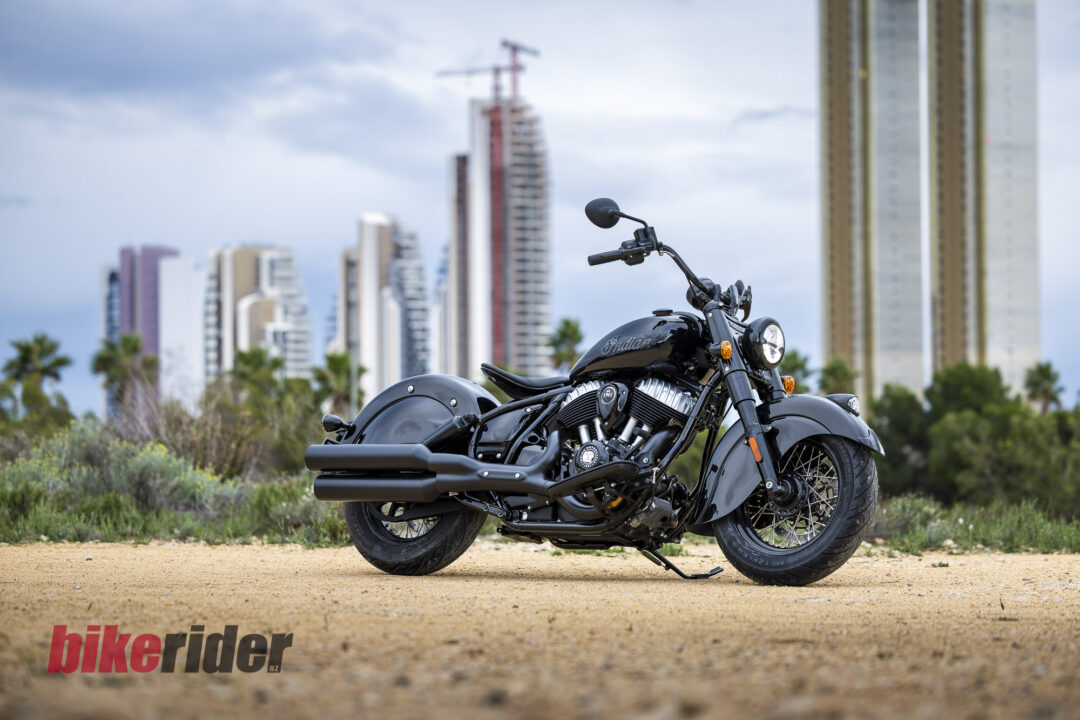 Black Indian motorcycle in the foreground on a dirt surface with a city skyline of high-rise buildings behind it.