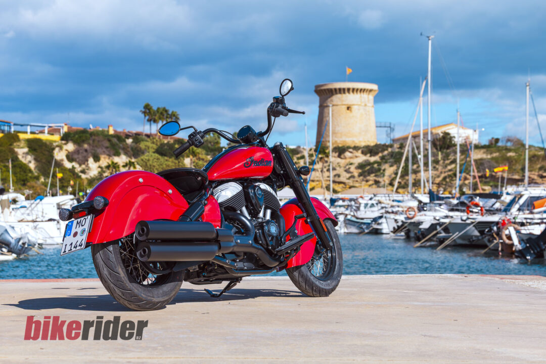 Red Indian motorcycle parked near a marina with boats in the background and a 'bikerider.nl' logo on the ground