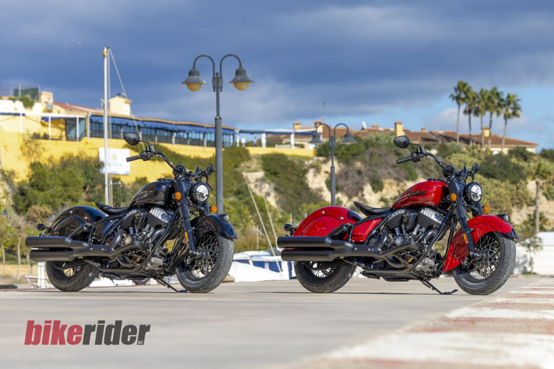 Two motorcycles parked side by side on a concrete surface with a waterfront town and palm trees in the distance, and a red 'bike rider' watermark at the bottom left.