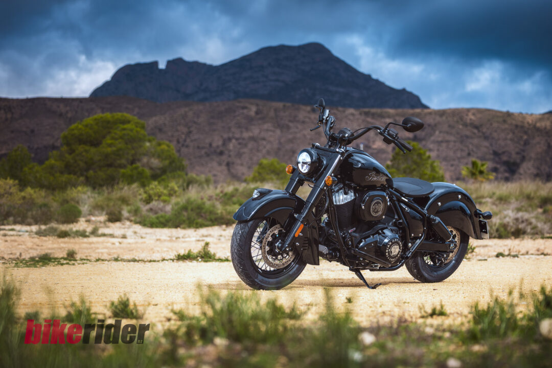 Black cruiser motorcycle parked on a sandy desert with rugged hills and a stormy sky in the background.