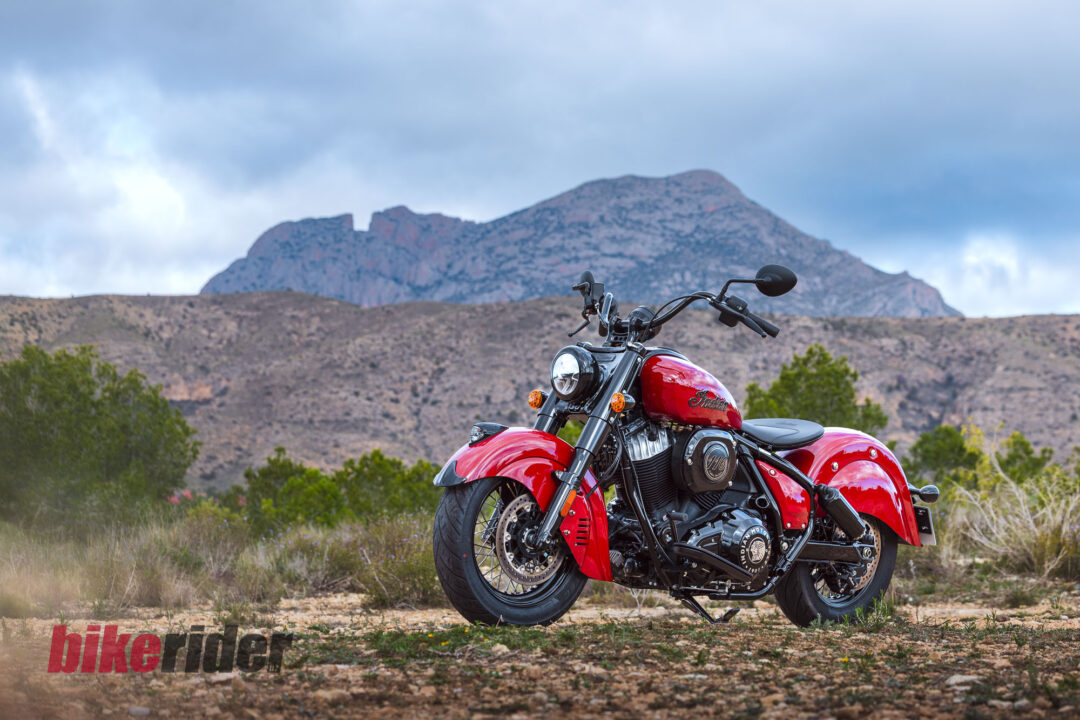 Red cruiser motorcycle in the foreground on a dirt path, desert hills in the background under a cloudy blue sky; watermark text 'bike rider' at lower edge.