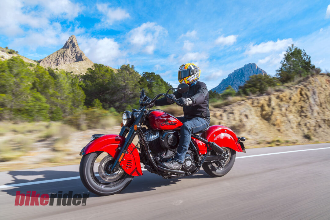 Motorcyclist wearing a colorful helmet and black gear riding a red Indian motorcycle along a highway with rocky hills and blue sky in the background.
