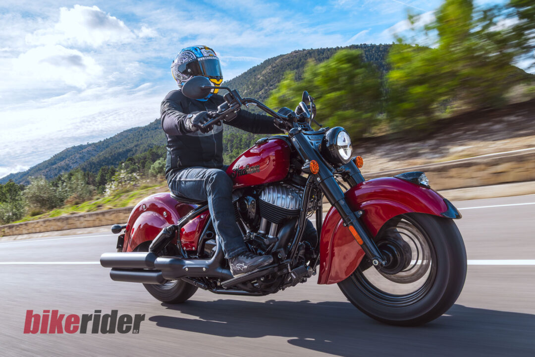 Rider wearing a helmet and black gear on a red Indian motorcycle zooms along a mountain highway with motion blur in the background.