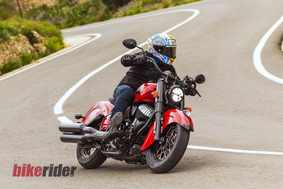 Motorcyclist in a colorful helmet leaning into a curve on a red motorcycle on a winding road with greenery nearby.