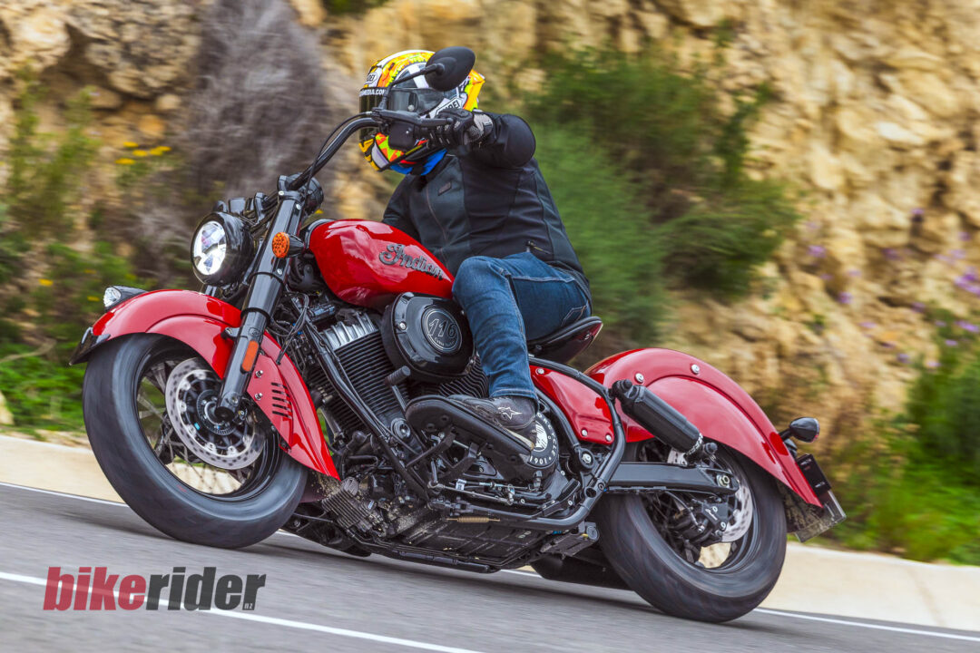 Motorcyclist in a colorful helmet and black gear leaned into a turn on a red Indian motorcycle on a road.