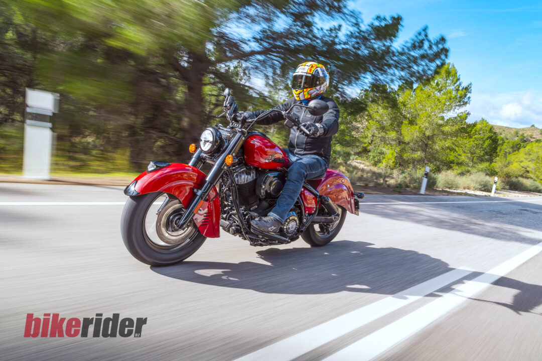 Motorcyclist wearing a colorful helmet rides a red motorcycle on a sunny open road with trees in the background.