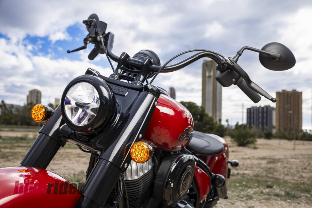 Close-up of a red and black motorcycle from the left, featuring a round headlight, orange turn signals, and chrome engine against a cloudy sky.