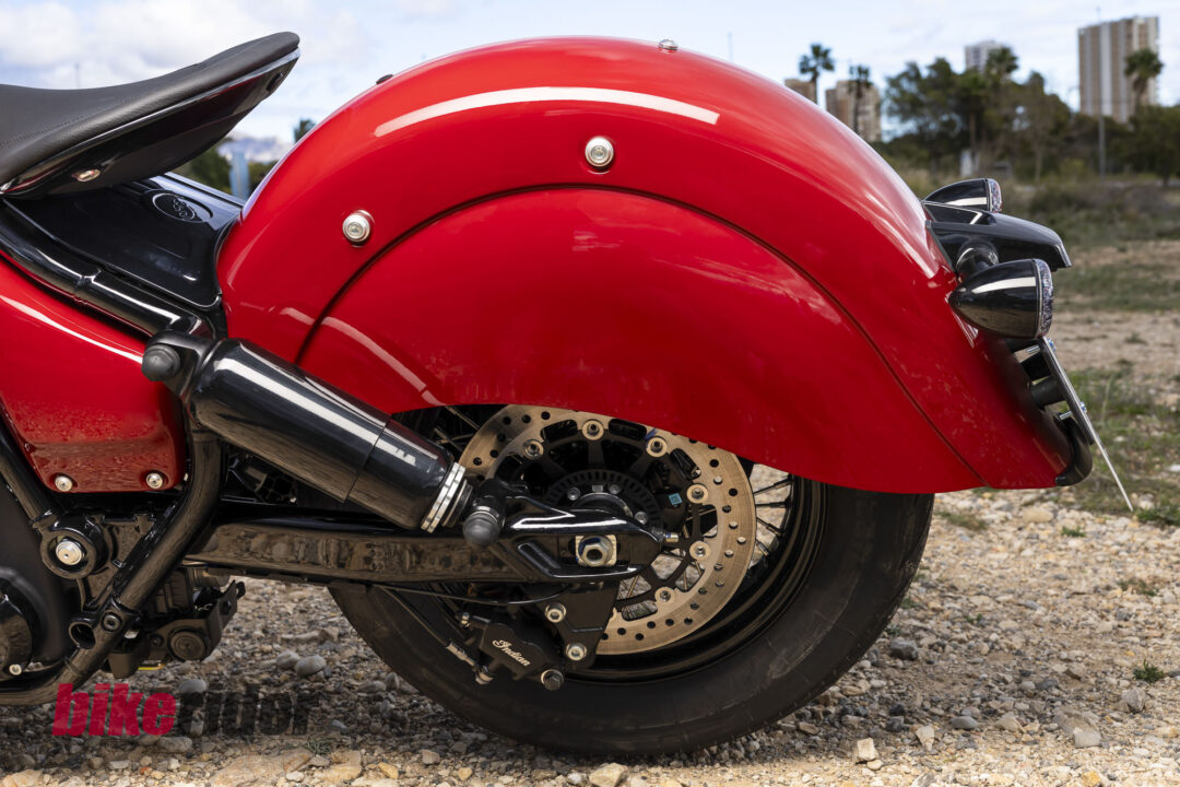 Close-up of a red motorcycle's rear section on a rocky ground, showing the curved fender, disc brake, and black exhaust components.