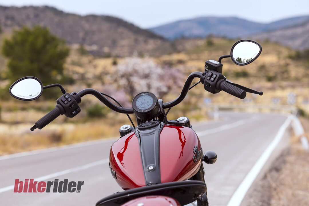 Close-up of a red motorcycle fuel tank and handlebars with mirrors and speedometer, on a scenic desert road.