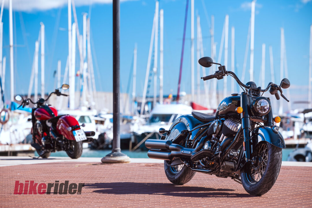 Close-up of a black motorcycle with chrome accents on a brick promenade, marina with sailboats blurred in the background.