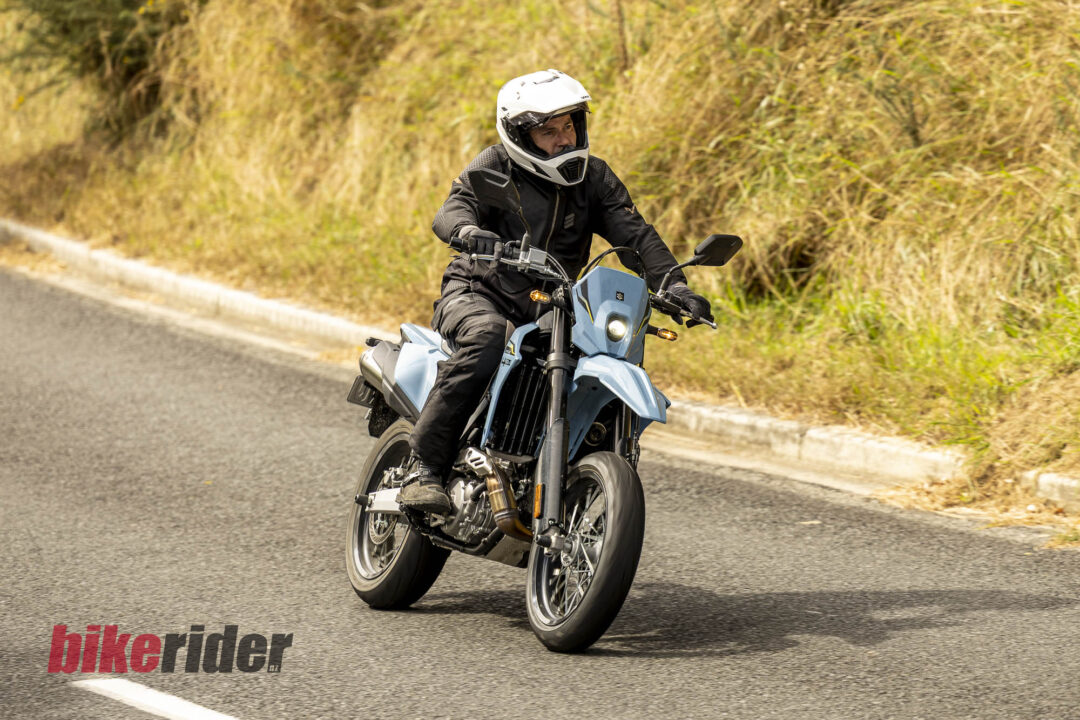 Motorcyclist in black gear and a white full-face helmet riding a blue motorcycle on a sunlit road with dry grass along the roadside.