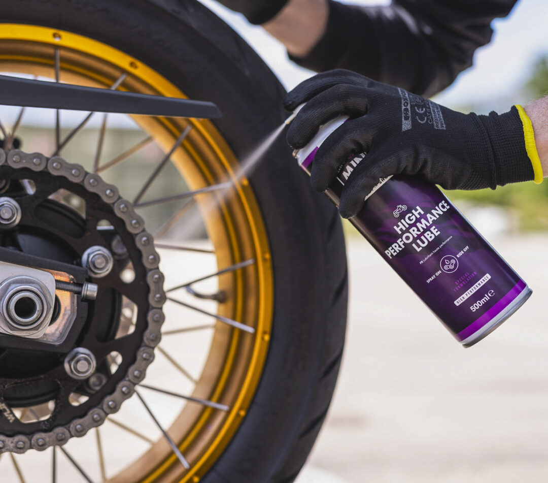 Gloved hand applying lubricant to a bicycle chain near a yellow rim, with a purple bottle labeled 'High Performance Lubricant' nearby
