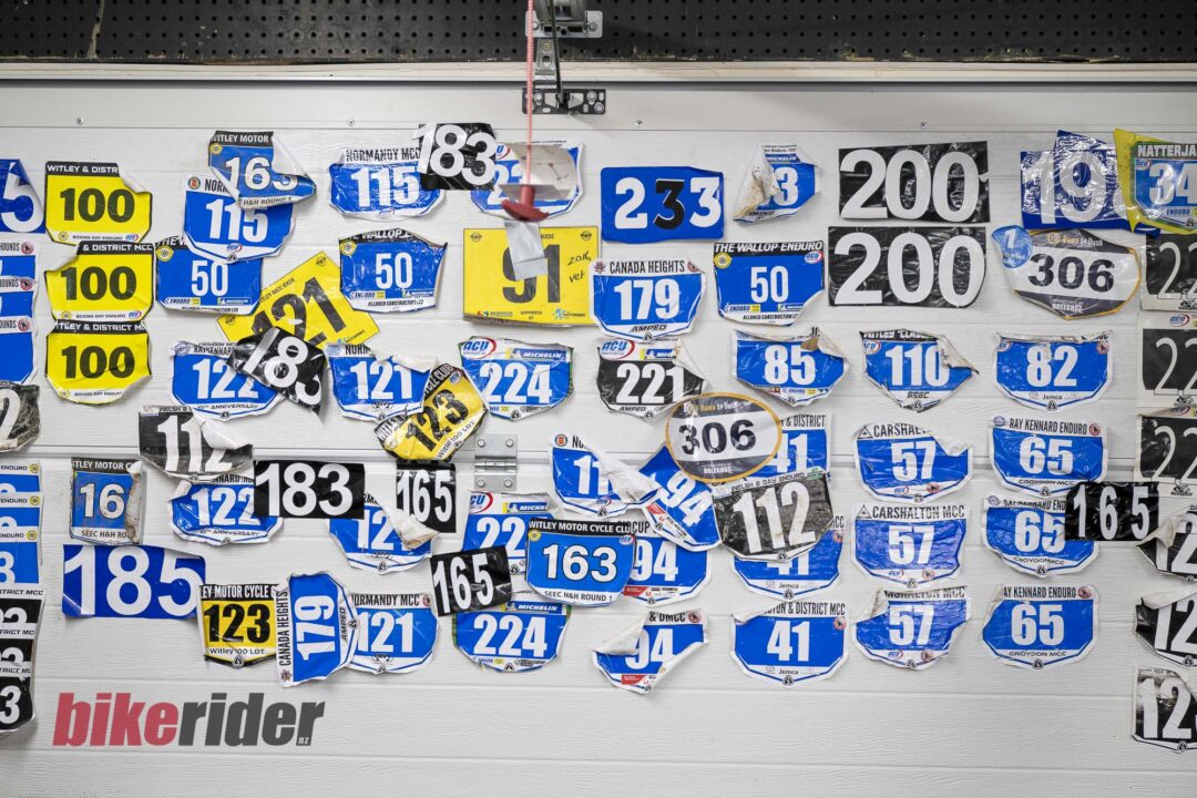20260617-_DSC3070-1080x720 Wall covered with overlapping bicycle race bibs in blue, yellow, and white; 'bike rider' logo visible at bottom.