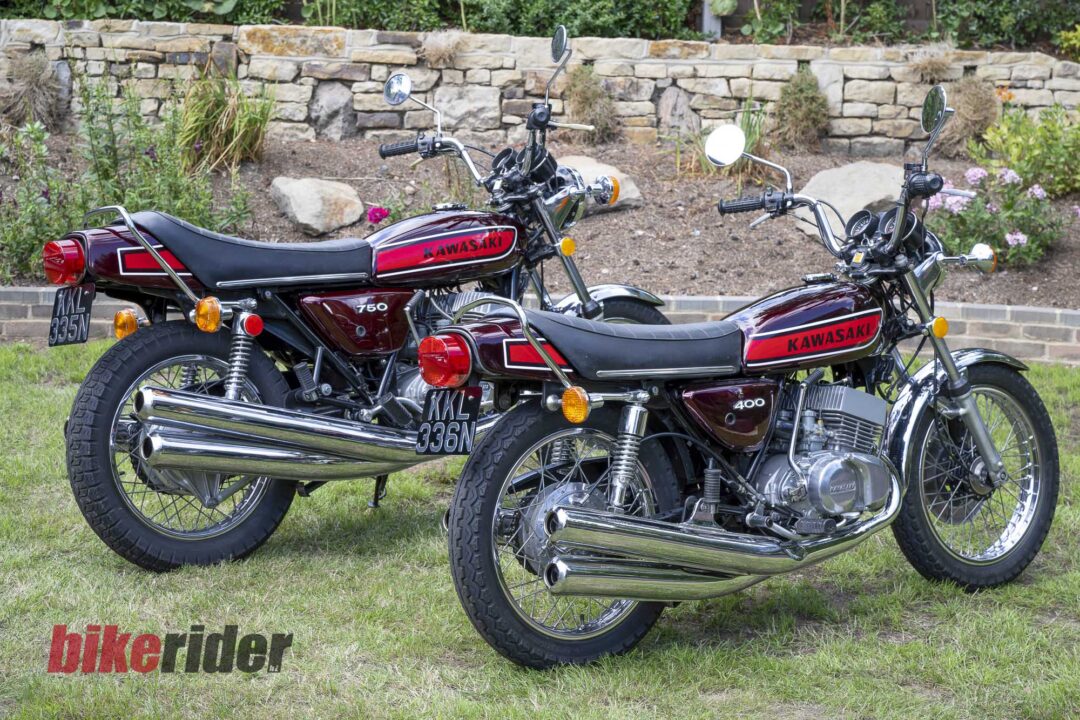 20260617-_DSC3332-1080x720 Two vintage Kawasaki motorcycles parked on a grassy area with a stone wall behind them.