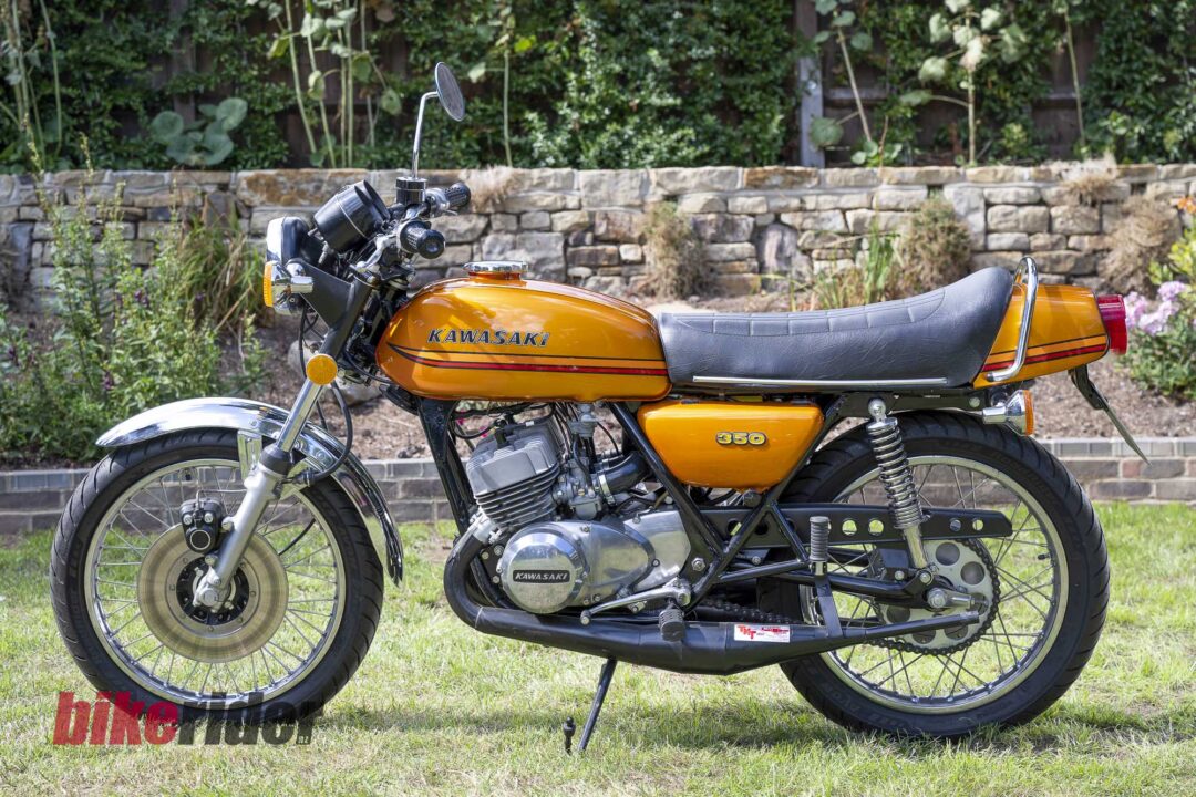 Orange Kawasaki motorcycle parked on grass with a stone wall in the background, showing the side profile and chrome details.
