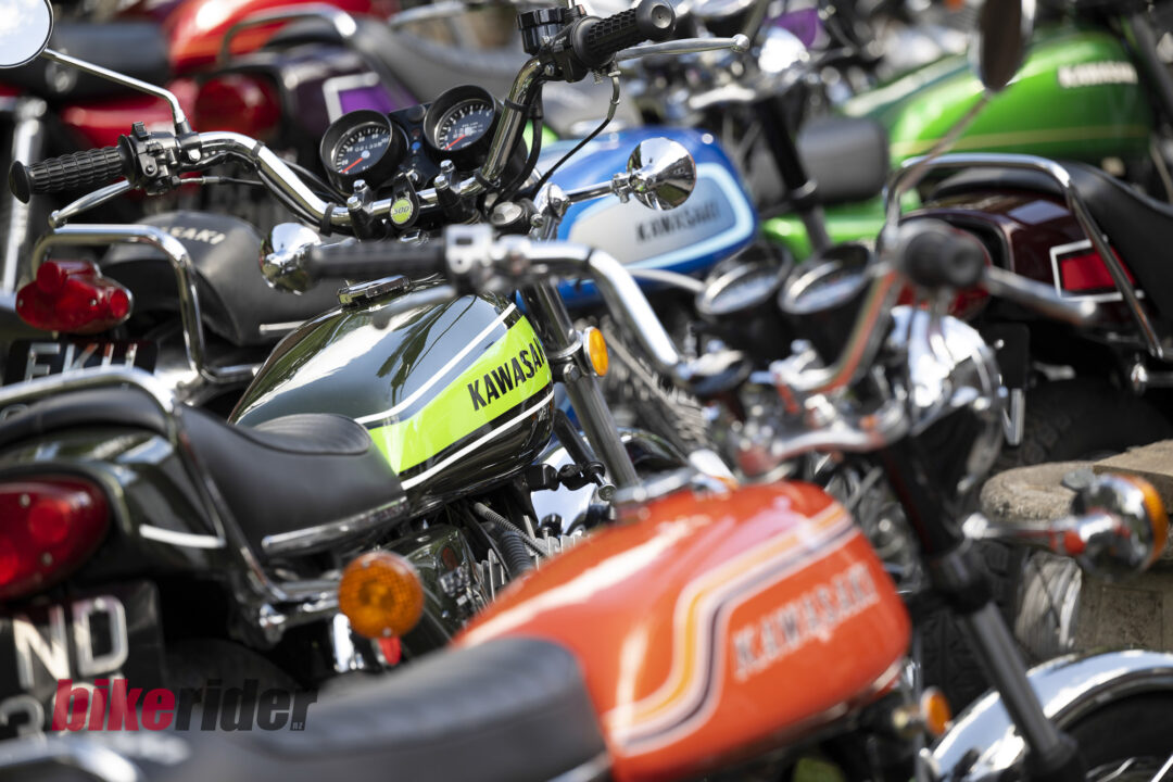 Close-up of several vintage motorcycles parked together, foreground green Kawasaki tank with chrome handlebars and gauges visible among colorful bikes behind.