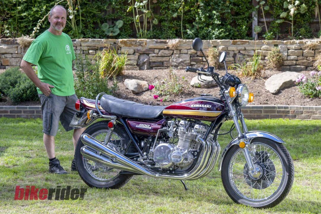 Man in a green shirt standing beside a red Kawasaki motorcycle in a sunny backyard garden with a stone wall behind.