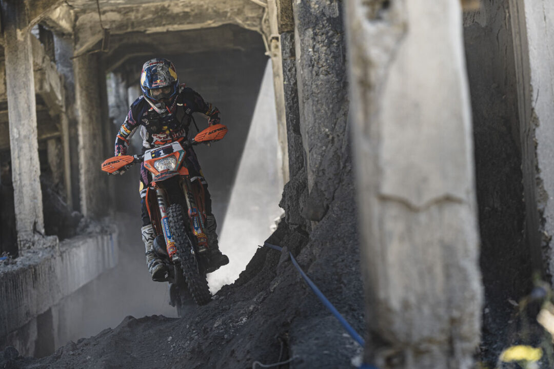 Dirt bike rider in full gear navigating a rocky tunnel, kicking up dust as they ride through an assault-ruin environment.
