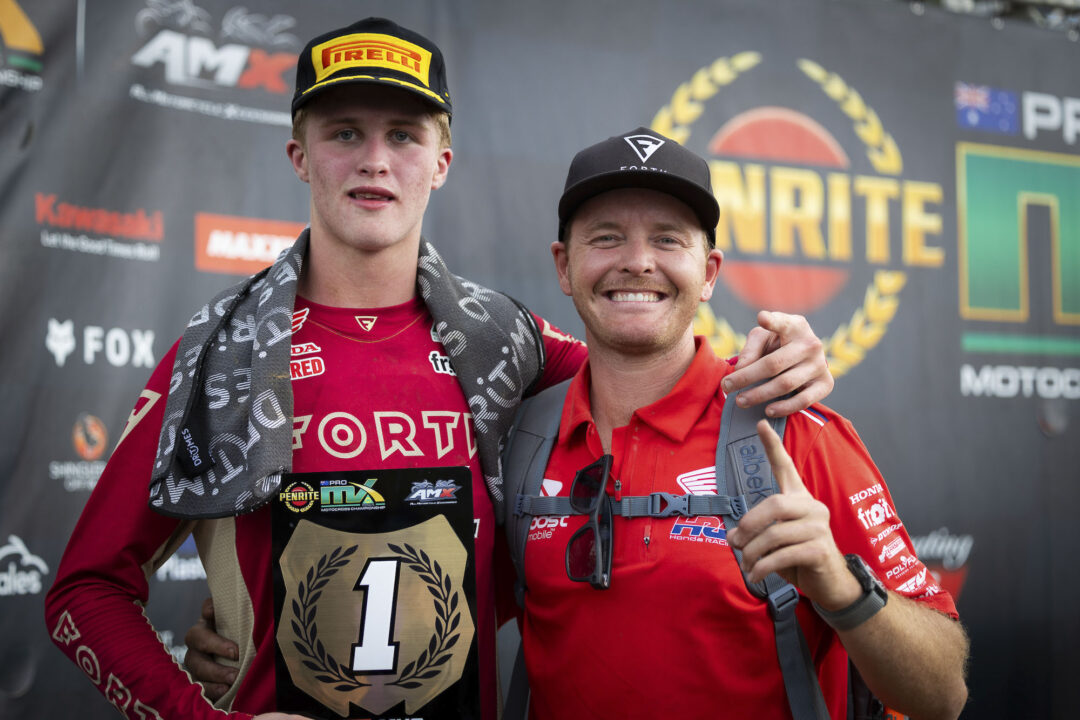 Two motocross racers pose after a podium win: left is a young rider in red with a Pirelli cap, holding a first-place plaque; right is a smiling teammate in red shirts.