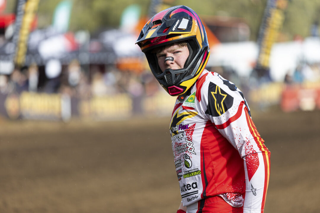 Motocross rider in red-and-white gear and a colorful full-face helmet on a dirt track with blurred spectators in the background.
