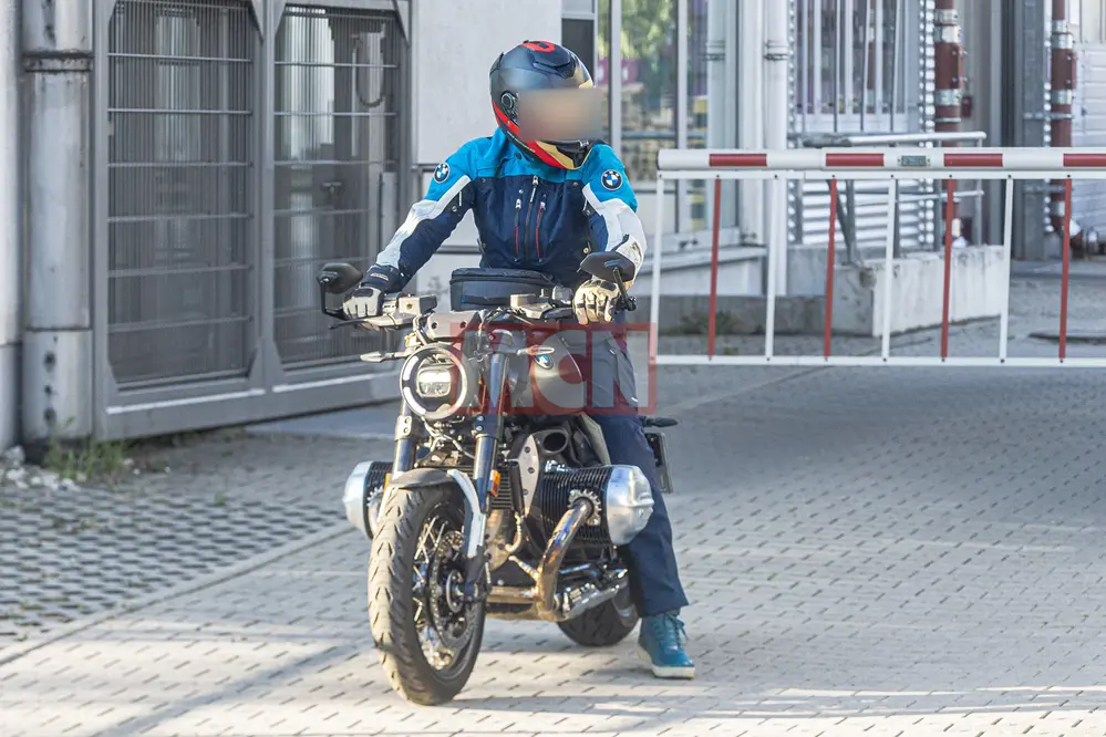 Motorcyclist in blue protective gear on a retro-style bike waiting at a striped barrier gate on a paved city street.