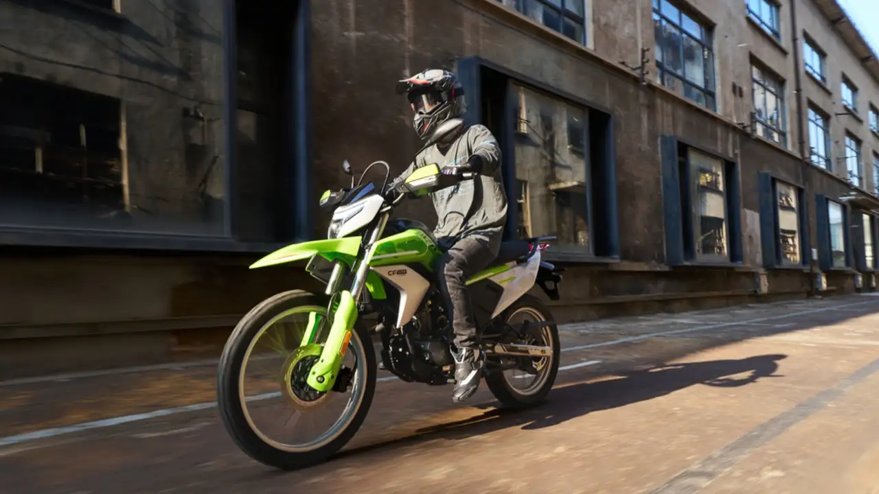 Motorcyclist wearing a helmet rides a bright green dirt bike on an urban street with industrial buildings in the background.
