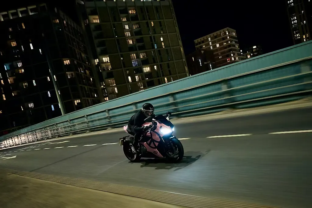 Motorcyclist in dark gear riding a pink-and-black sport bike at night on an city highway, illuminated by the bike's headlight with tall buildings in the background.