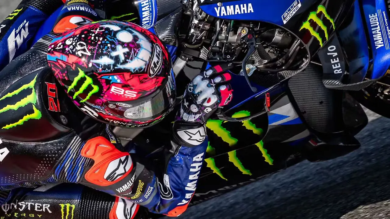 MotoGP rider leaning into a turn on a blue Yamaha bike, helmet with pink/blue artwork.