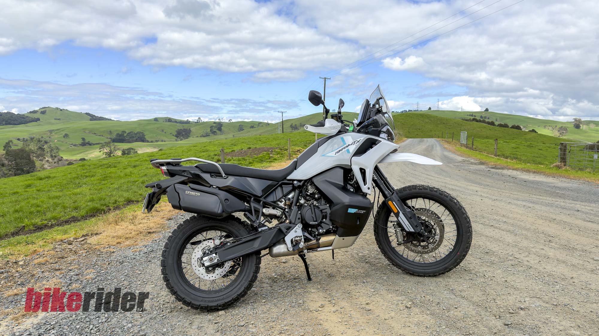 White CFMOTO 1000MT-X adventure bike parked on a gravel road among green hills
