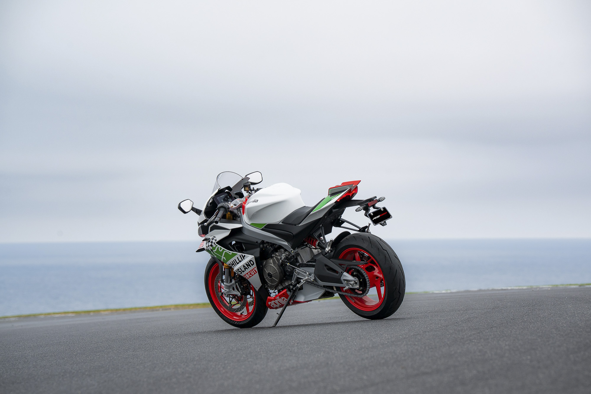 Aprilia RS 660 Phillip Island Tribute viewed from the rear left, showing its red wheels beside the ocean.