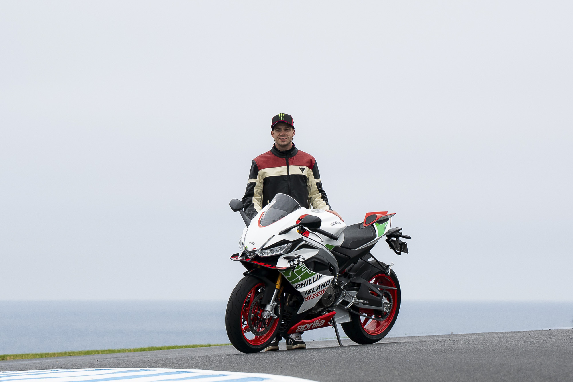 Marco Bezzecchi standing behind the Aprilia RS 660 Phillip Island Tribute beside the track and ocean.